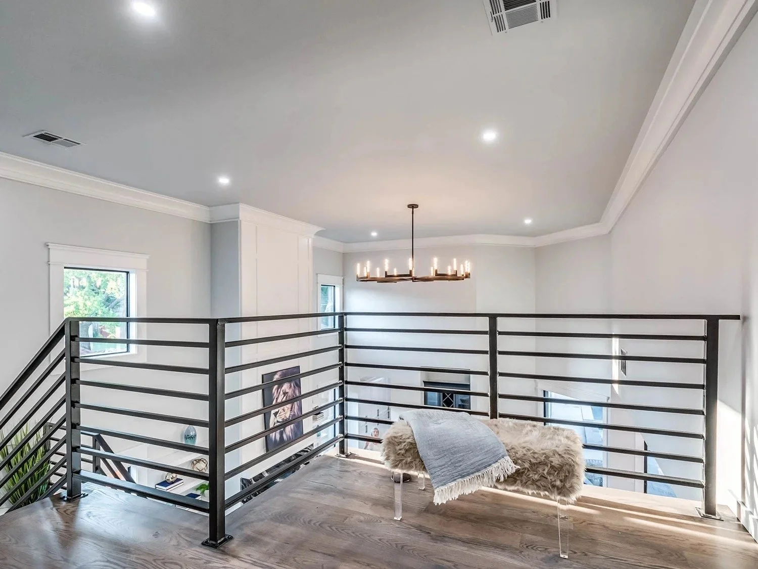 Upper floor interior with metal railing, wood flooring, a white cozy chair with a blanket, a modern chandelier, and large windows in a bright, modern home.