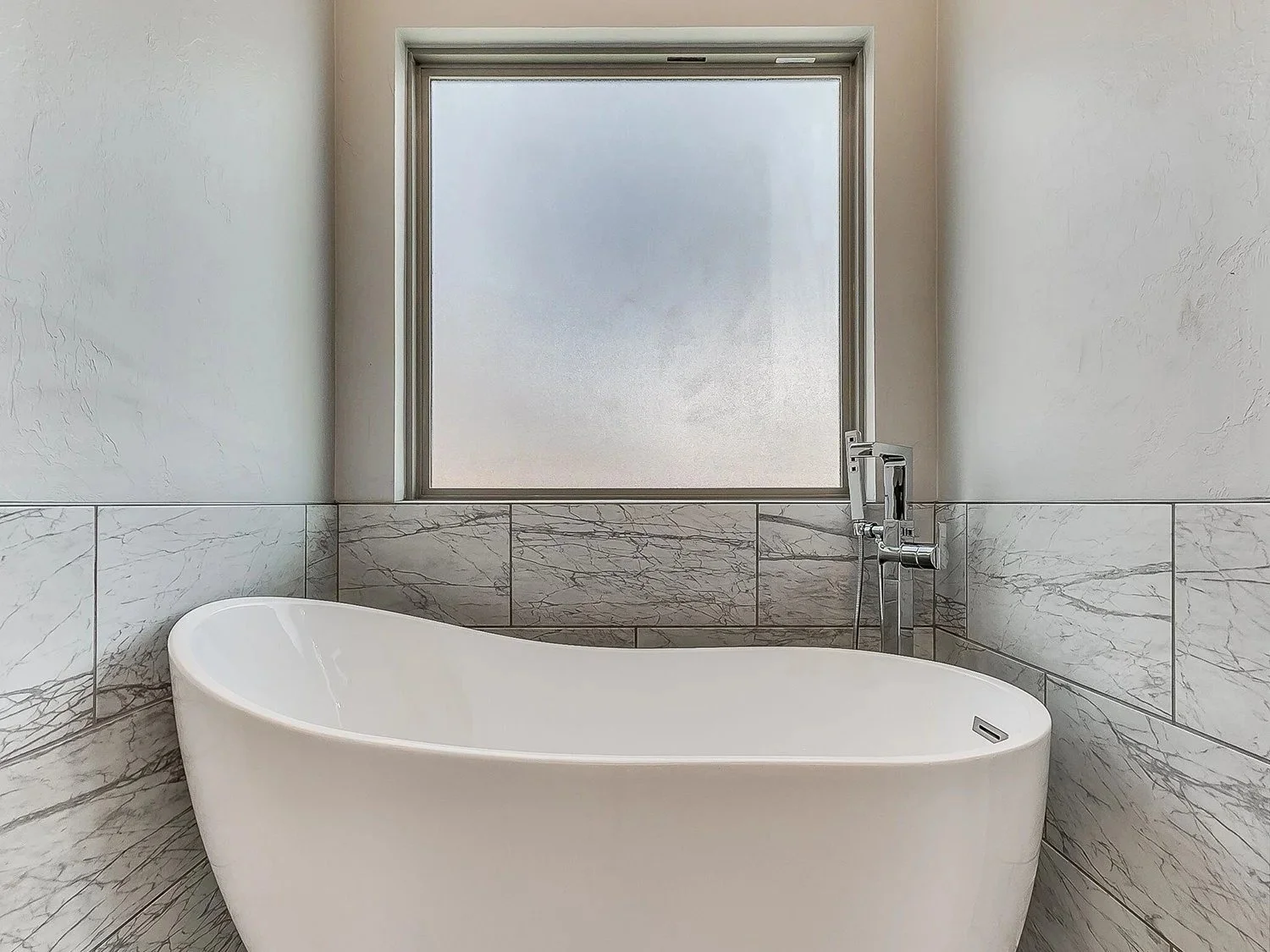 Modern bathroom with a freestanding bathtub, marble tiles, and frosted window.