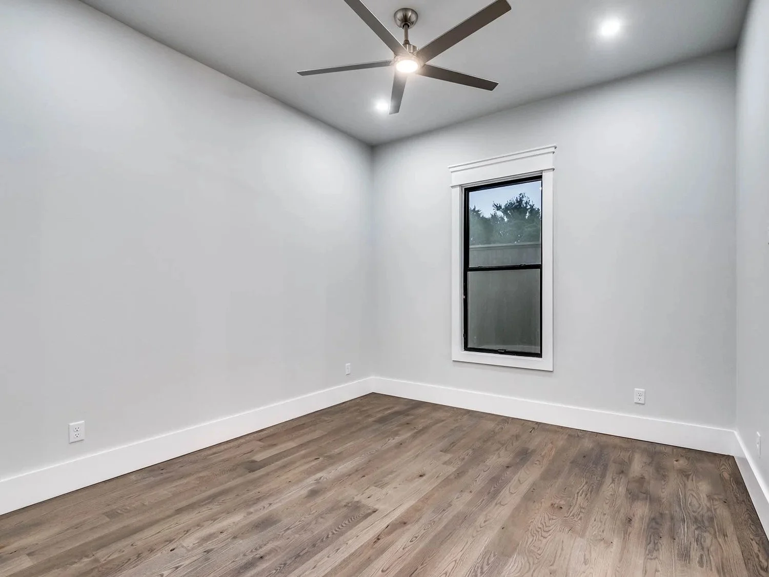 Empty room with white walls, a window, a ceiling fan, hardwood floor, and electrical outlets.