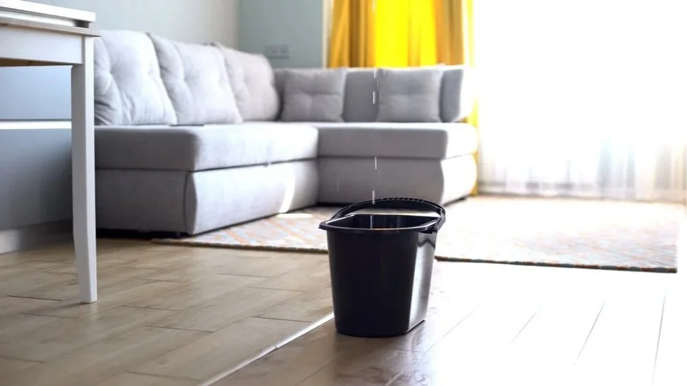 A living room with light hardwood floors, a gray sofa, a small white side table, and yellow curtains. There is a black trash can in the center of the room.