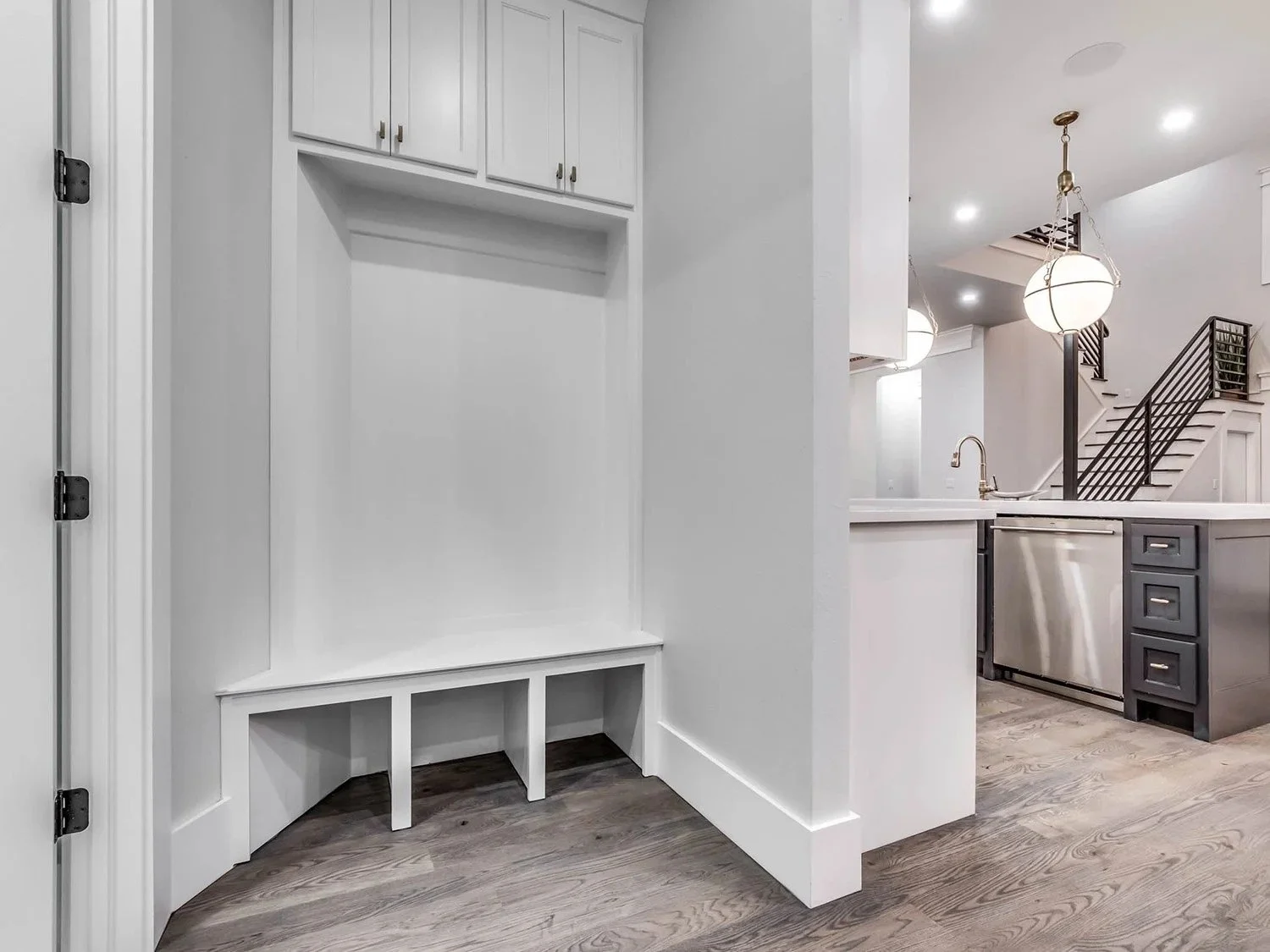 Empty white built-in closet with bench and upper cabinets in a modern kitchen with dark lower cabinets, stainless steel appliances, and a staircase with black railings in the background.