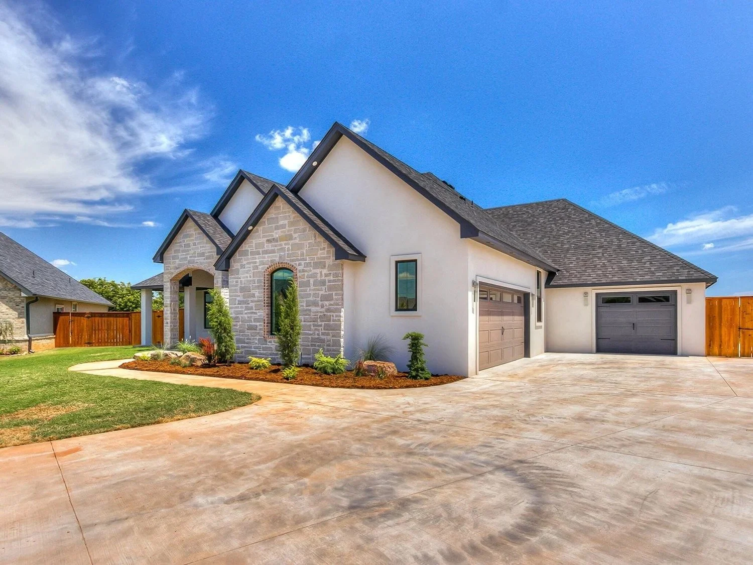 Modern house with stone and white exterior, two garage doors, landscaped front yard, concrete driveway, wooden fence, under a blue sky with clouds
