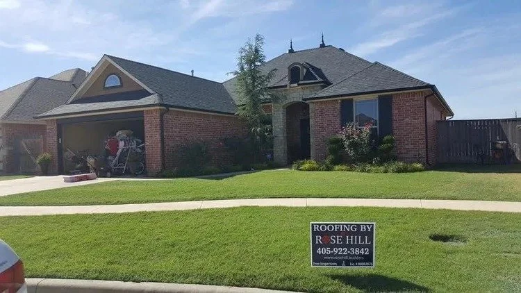 A brick house with a covered garage and landscaped yard. There is a sign in the lawn that reads "Roofing by Rose Hill" with a phone number.