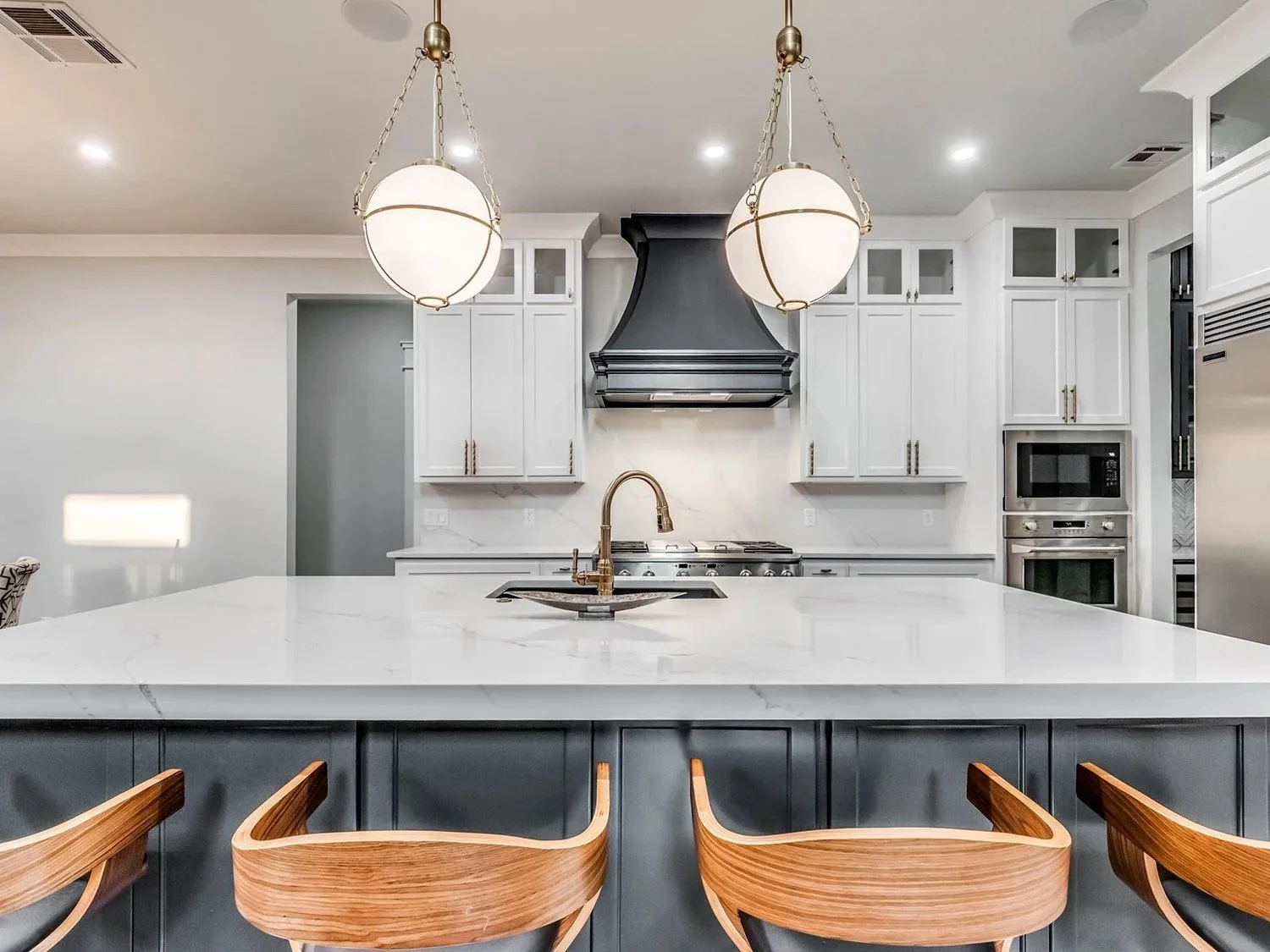 Modern kitchen with white marble countertops, black cabinetry, and a large island. Features stainless steel appliances, a black range hood, and wooden chairs.
