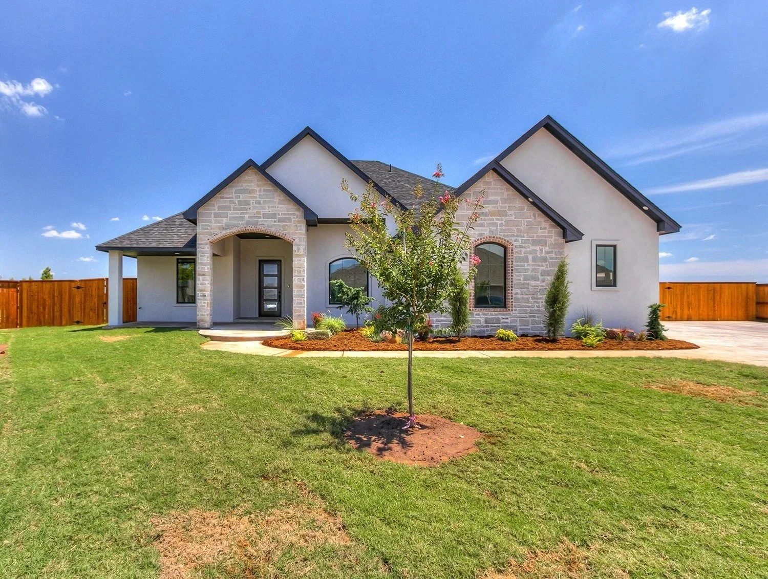 Modern house with stone and stucco exterior, front yard with green grass and small trees, blue sky with clouds.