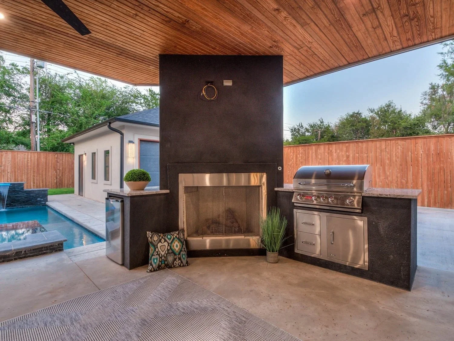 Outdoor kitchen with built-in stainless steel grill, black fireplace, and swimming pool in backyard surrounded by wooden fence and trees