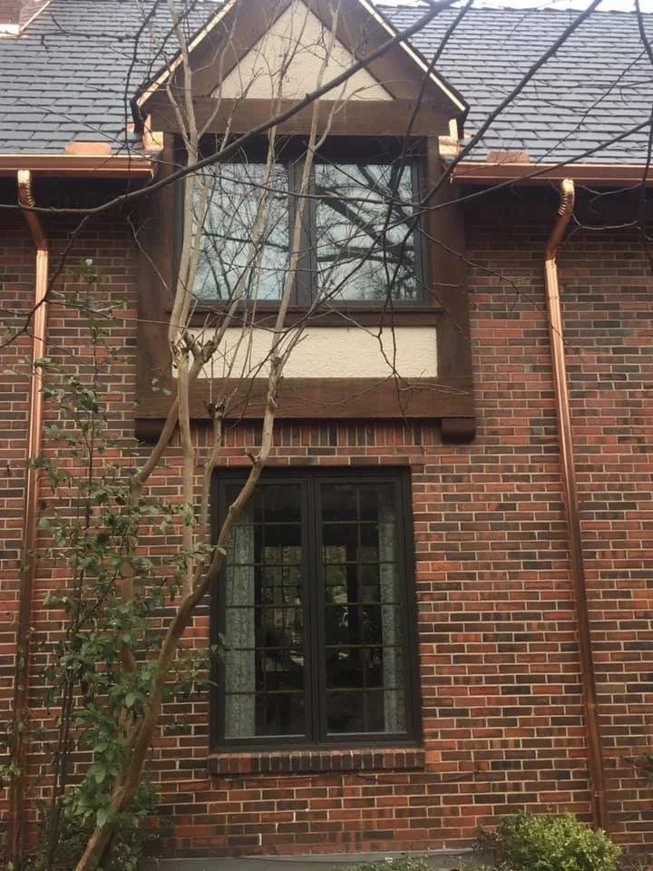 A brick house with black-framed windows, some of which have grids. The house has a gabled roof with gray shingles. There are leafless tree branches in front of the windows, and copper-colored downspouts run down the sides of the house.