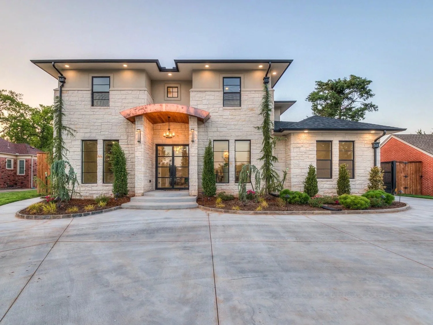 Modern two-story house with white stone exterior, large black windows, and a driveway. Front yard has landscaping with trees and shrubs.