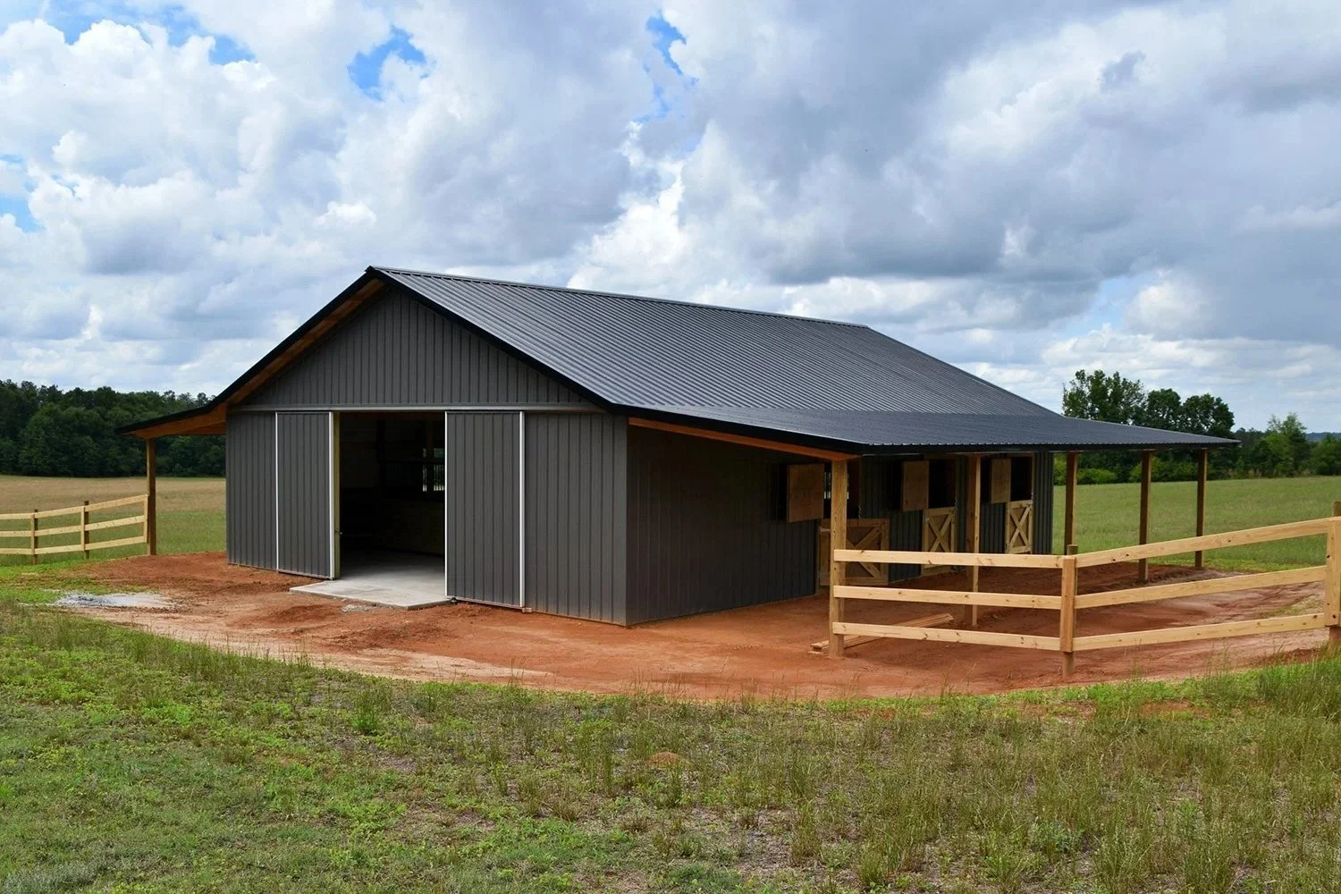 A newly built black metal barn with sliding doors, situated in a grassy field with a dirt area in front, surrounded by a wooden fence, under a partly cloudy sky.