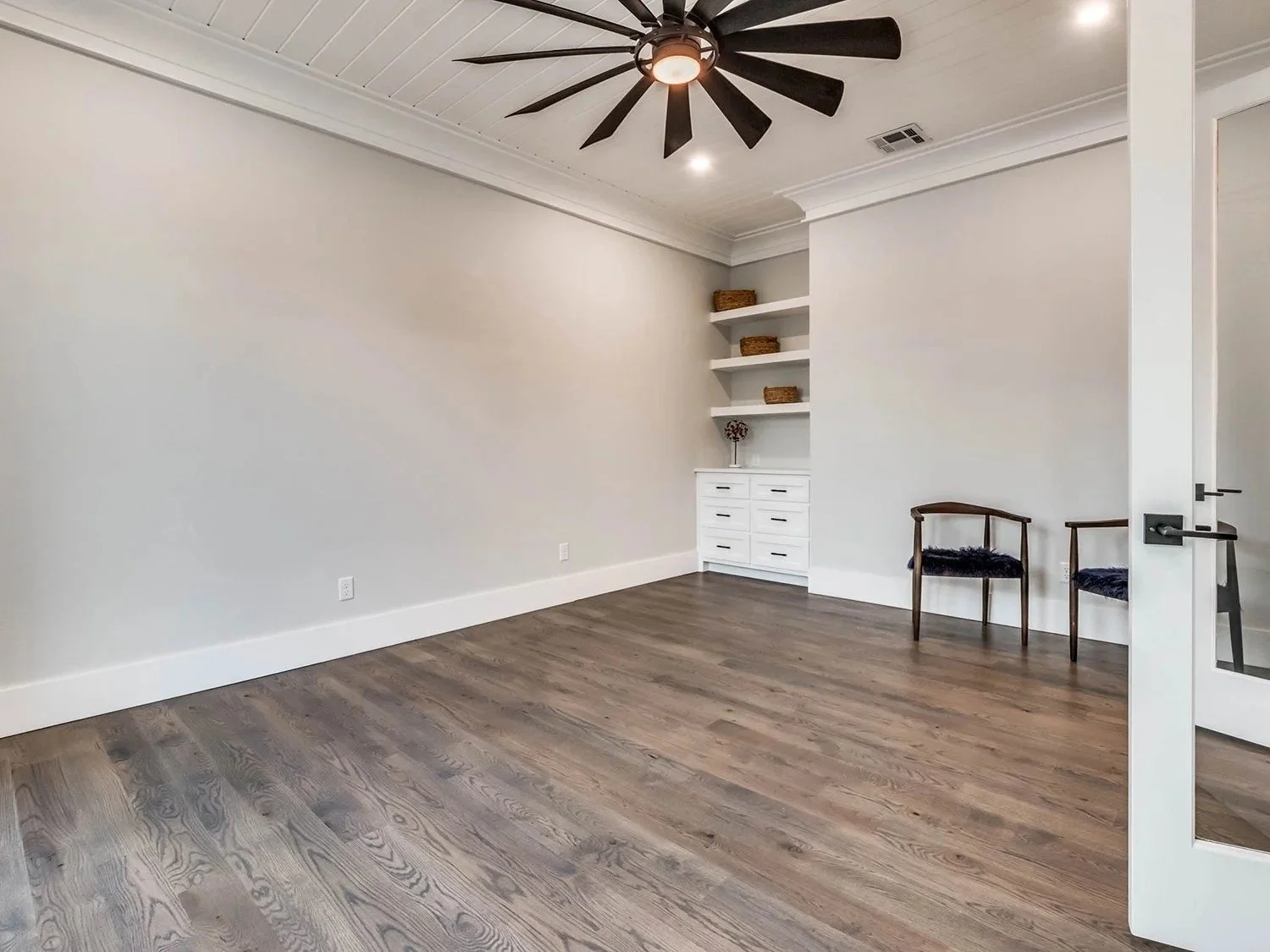 A spacious, empty room with white walls, hardwood floor, a black ceiling fan, and a white ceiling with recessed lighting. There are two black chairs with wooden frames in the corner and white built-in shelves with wicker baskets.