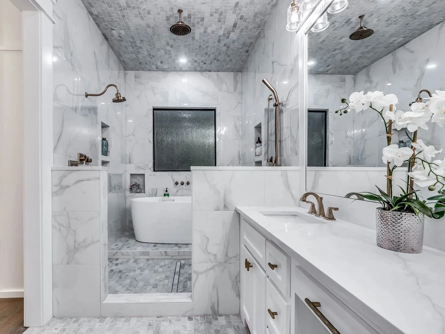 A modern bathroom with white Marble walls and a walk-in shower with a frosted window, a bathtub, and a vanity with a white countertop, gold fixtures, and a potted orchid plant.
