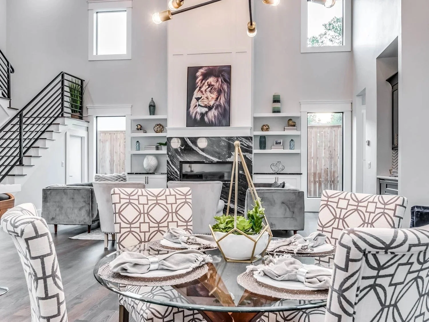 Bright living room with high ceilings, an artsy fireplace, and built-in shelves. A round dining table with patterned chairs, table settings, and a centerpiece with plants. Large windows let in natural light.