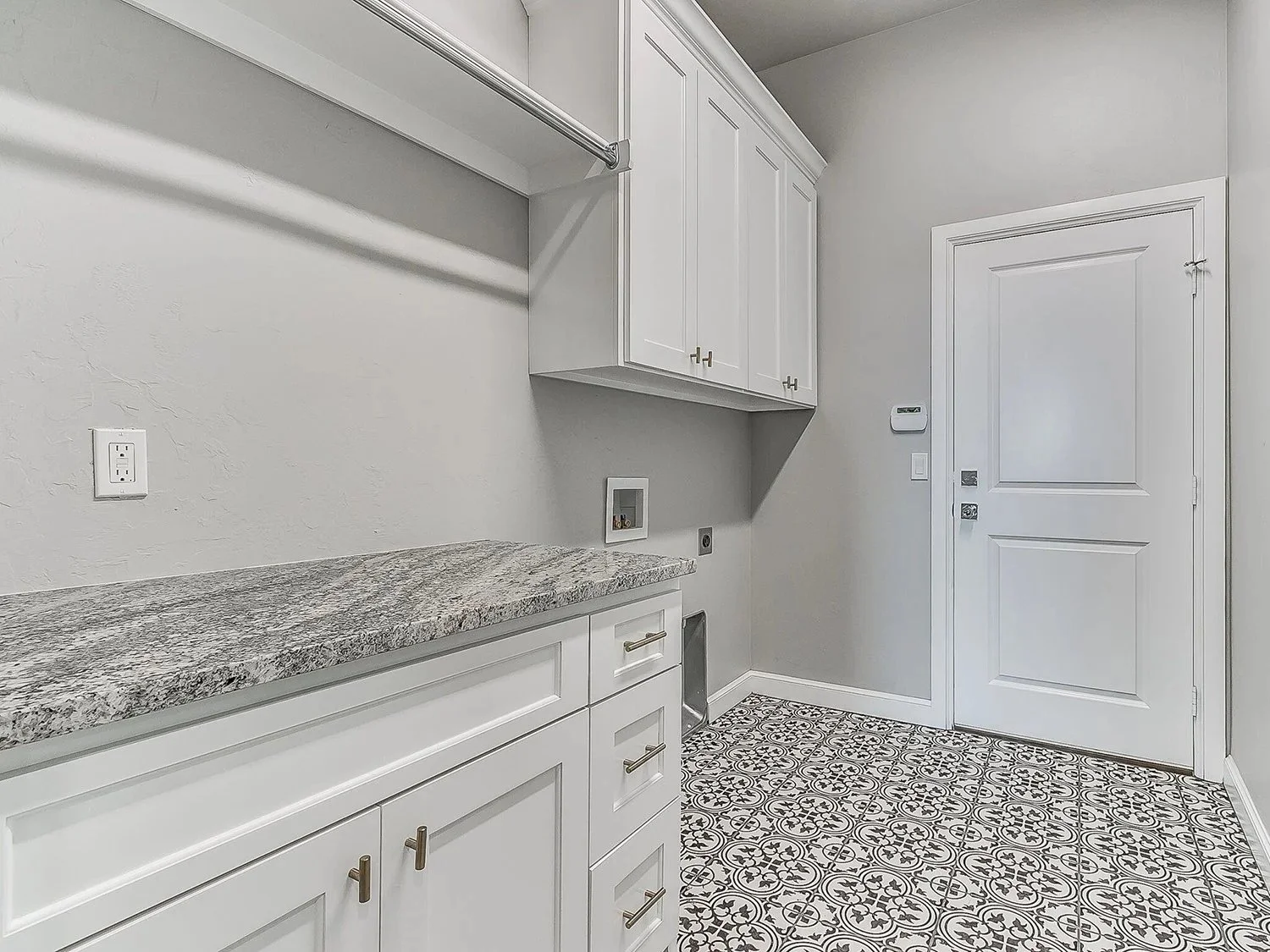 Empty laundry room with white cabinets, granite countertop, patterned floor tiles, and a closed door.