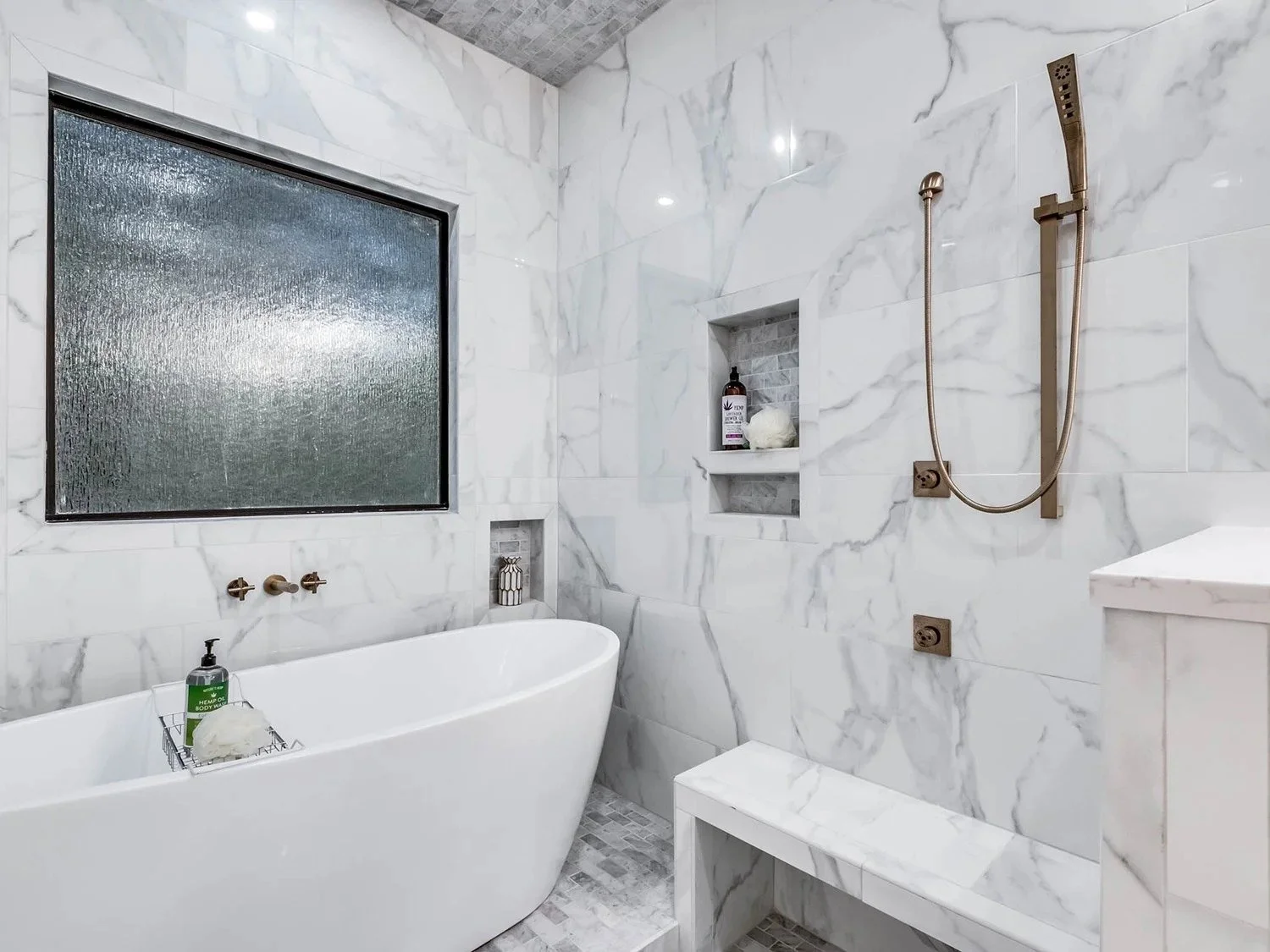 Modern bathroom with white marble walls, a freestanding bathtub, a frosted glass window, built-in shelves holding toiletries, and a walk-in shower with a brass rainfall showerhead.