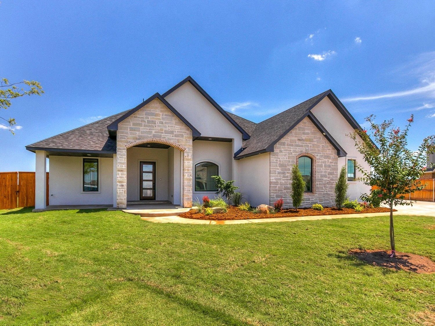 A modern suburban house with light-colored stone and stucco exterior, a dark shingled roof, a wooden front door, a small landscaped front yard, and a blue sky in the background.