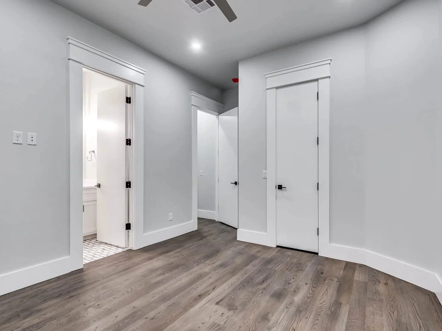 Empty room with white walls, white trim, hardwood floor, three closed white doors with black door handles, and a ceiling fan with a light.