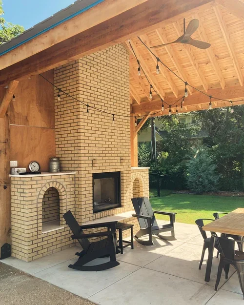 Outdoor patio with a brick fireplace, two black Adirondack chairs, a wooden dining table with chairs, string lights, a ceiling fan, and a grassy backyard with trees.