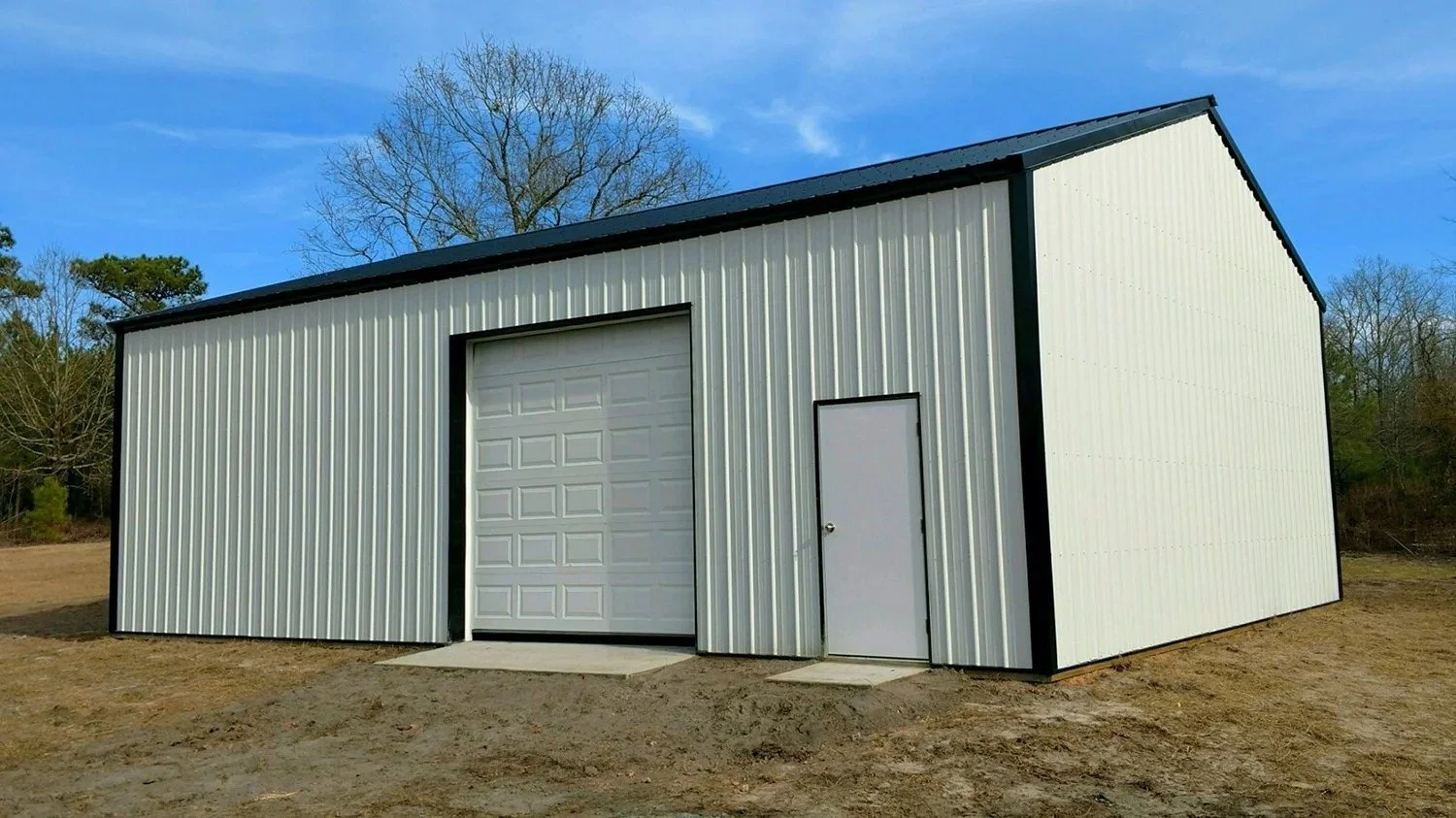 A metal storage building with a large garage door and a small door on a dirt lot, with trees and a blue sky in the background.