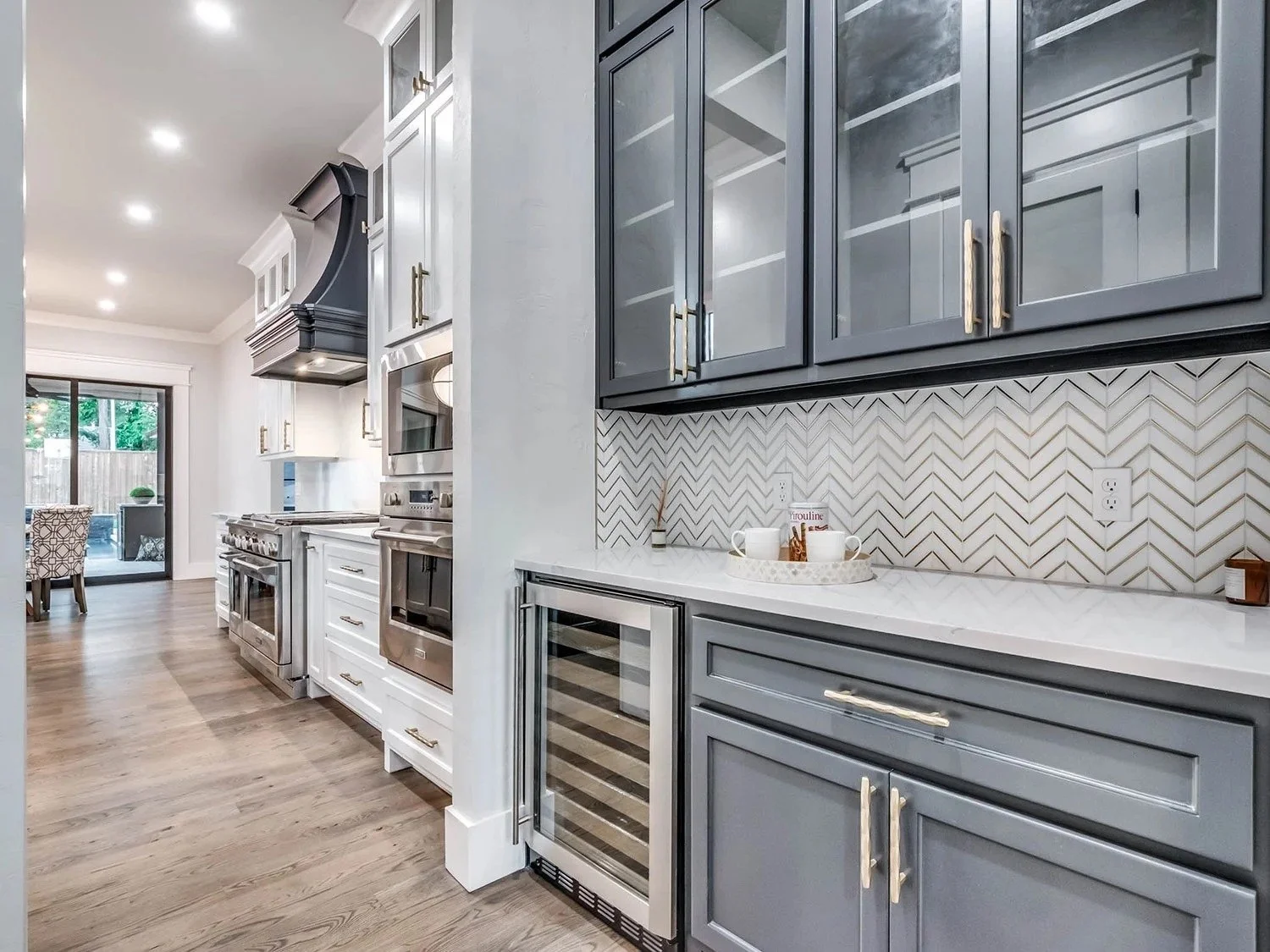 Modern kitchen with gray and white cabinets, white marble countertop, herringbone tile backsplash, stainless steel appliances, and hardwood flooring.