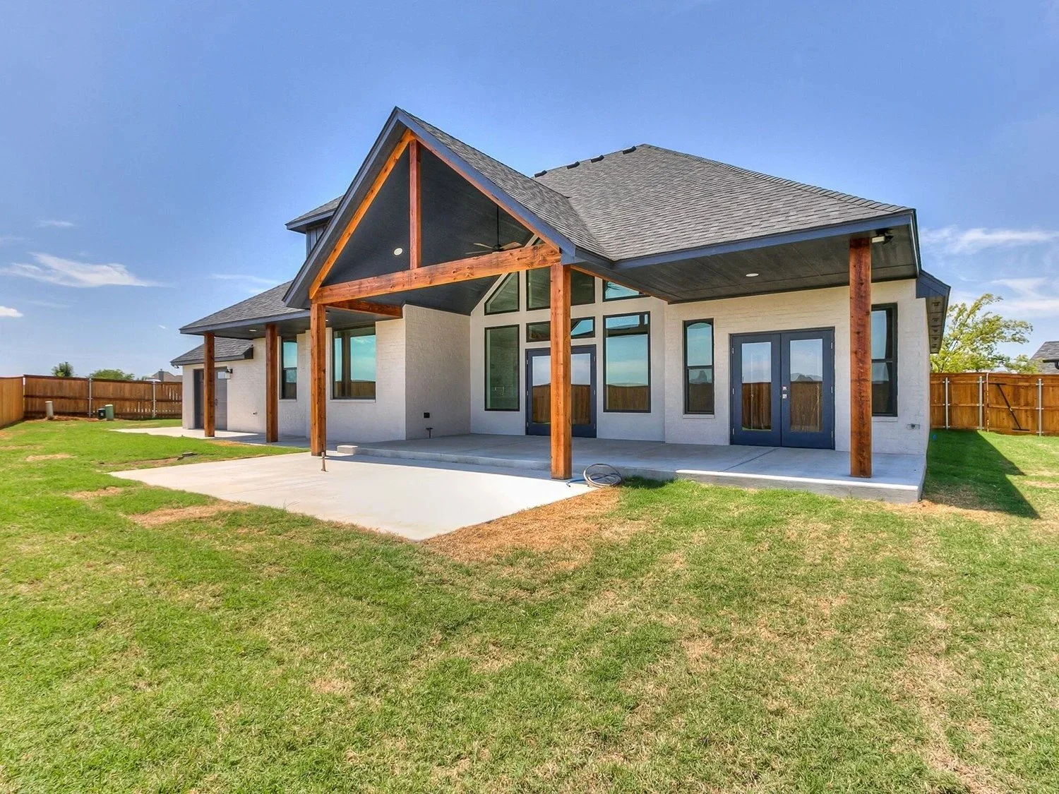 Brand new house with white brick exterior, large windows, and a covered porch with wooden beams, situated in a spacious backyard with green lawn and a wooden fence.