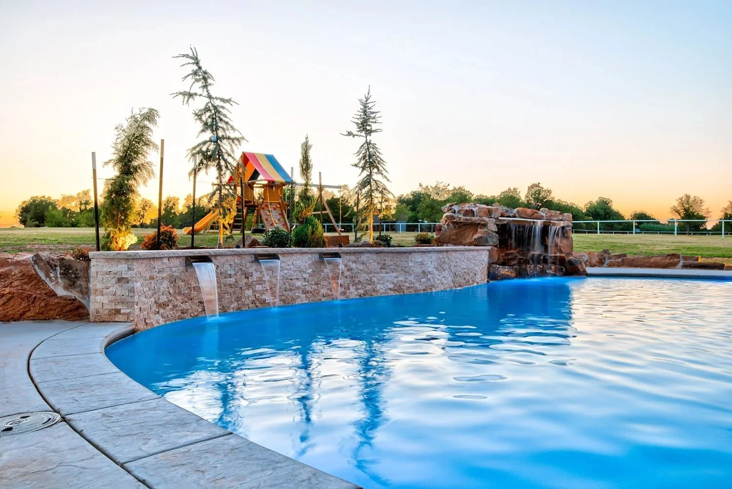 A backyard swimming pool with a rock waterfall feature, landscape with trees, a toy playset with a slide, and a sunset sky in the background.