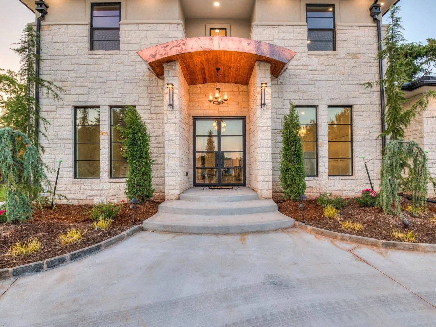 Front view of a modern house entrance with stone facade, glass double doors, curved steps, tall trees, and landscaped flower beds.