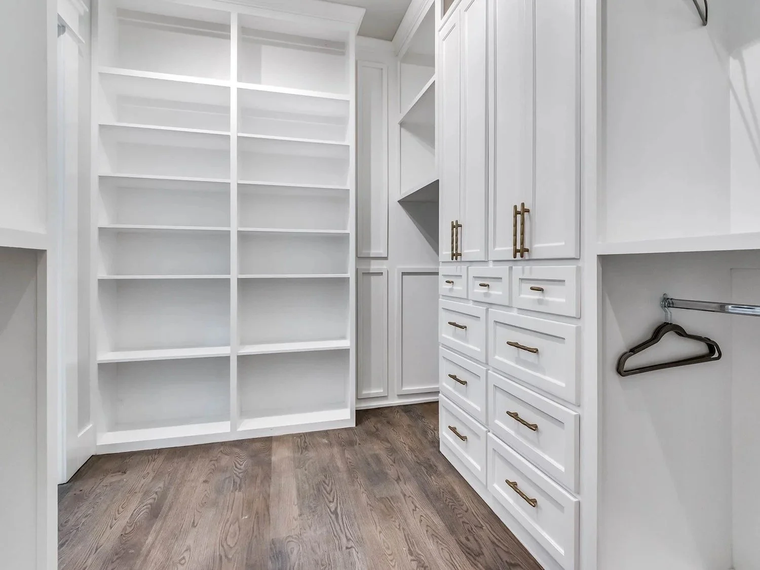 Empty walk-in closet with white built-in shelves, drawers, and hanging rods, and wood flooring.