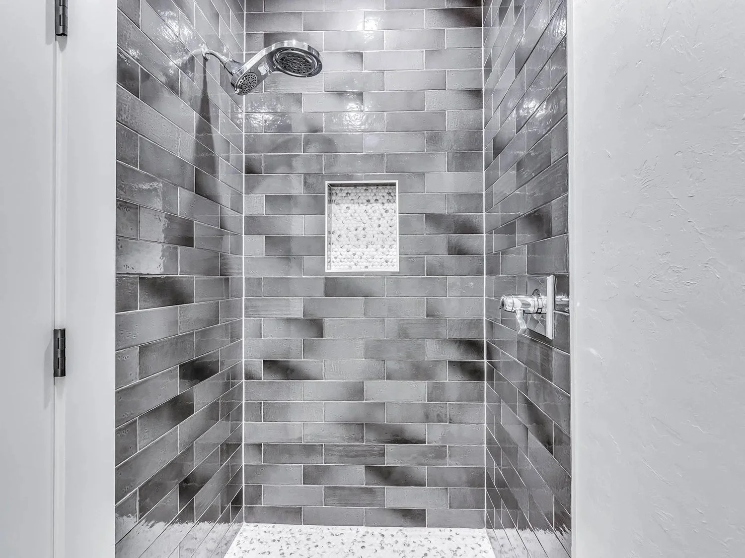 A modern shower with gray subway tile walls, a small niche with mosaic tiles, a chrome showerhead, and a textured white floor.