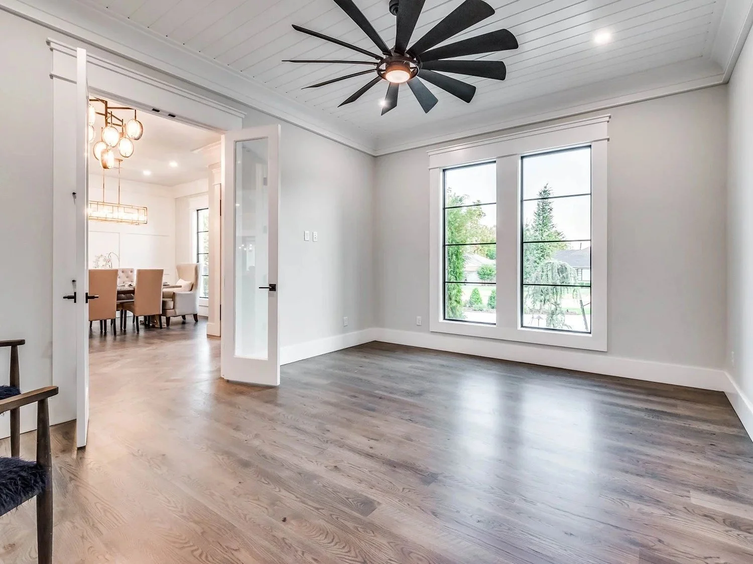 Empty living room with large windows, white walls, wood flooring, and a modern black ceiling fan.