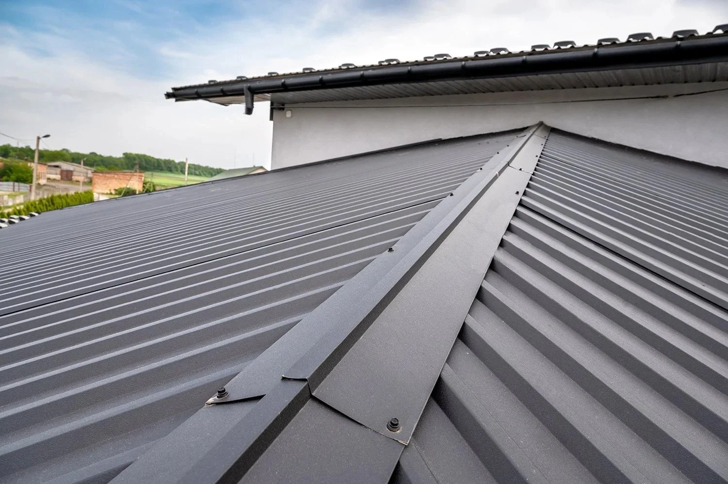 Close-up of a gray metal roof with ridges and a central ridge cap, with a building wall and cloudy sky in the background.