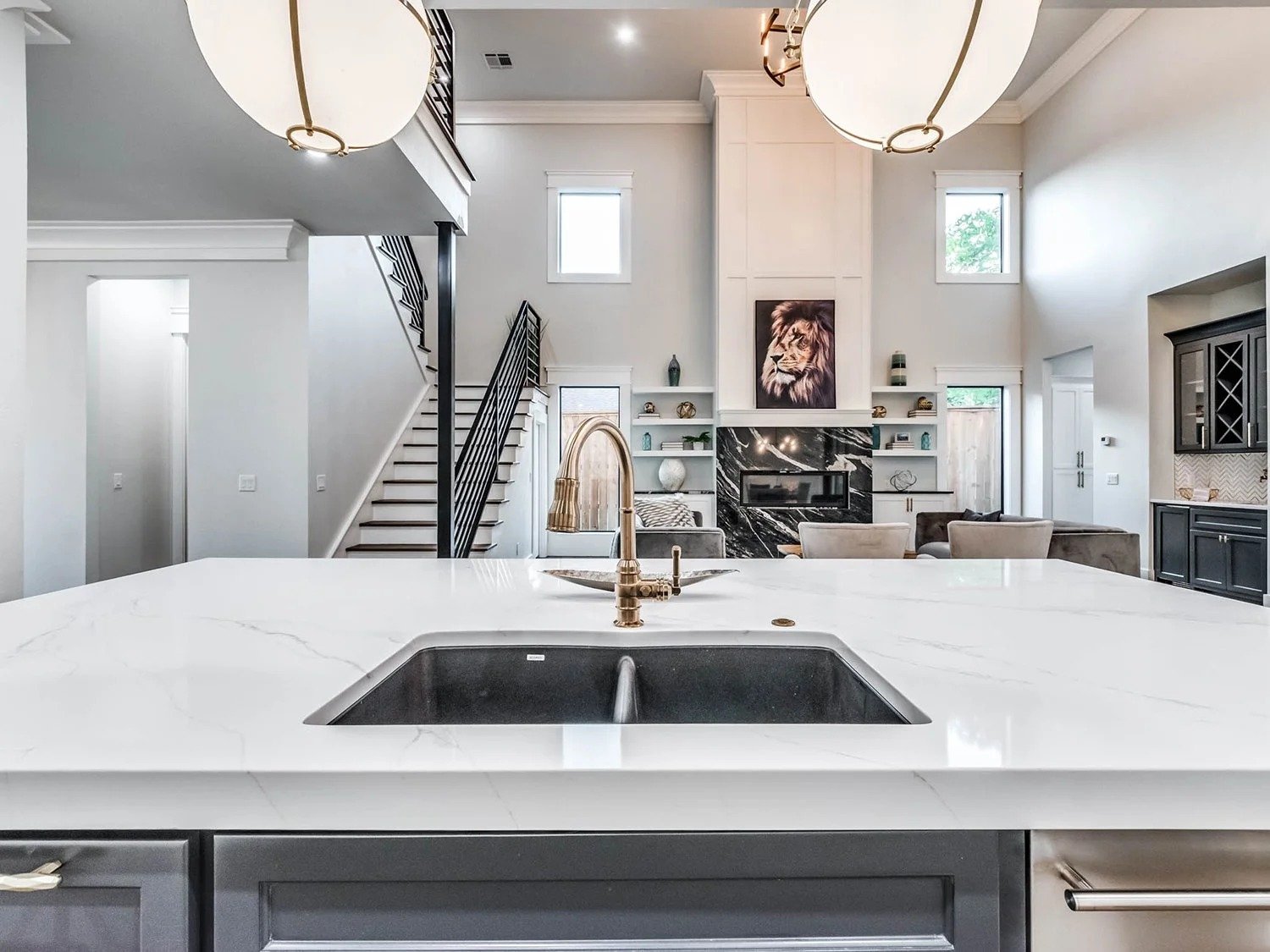 Modern kitchen with white marble countertops, a gold faucet, black sink, and a view of a living room with a fireplace, built-in shelves, and a lion painting above the fireplace.