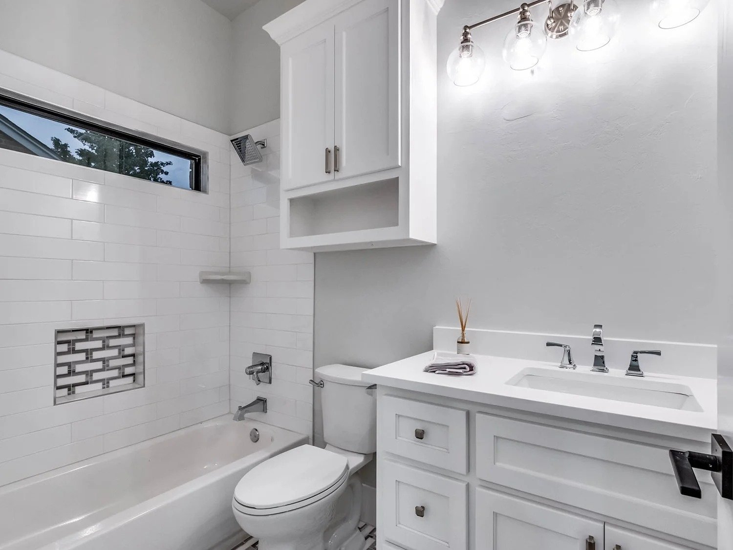 A modern white bathroom with a bathtub, a white toilet, a white vanity with a silver faucet, a large mirror, and a wall-mounted cabinet. There is a window above the bathtub, and a small shelf inside the shower area. The lighting fixture above the mir
