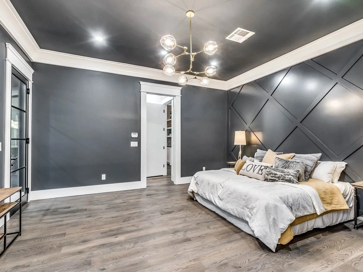 Modern bedroom with dark gray walls, a patterned accent wall, a bed with neutral and yellow pillows, a side table with a lamp, and a ceiling light fixture.