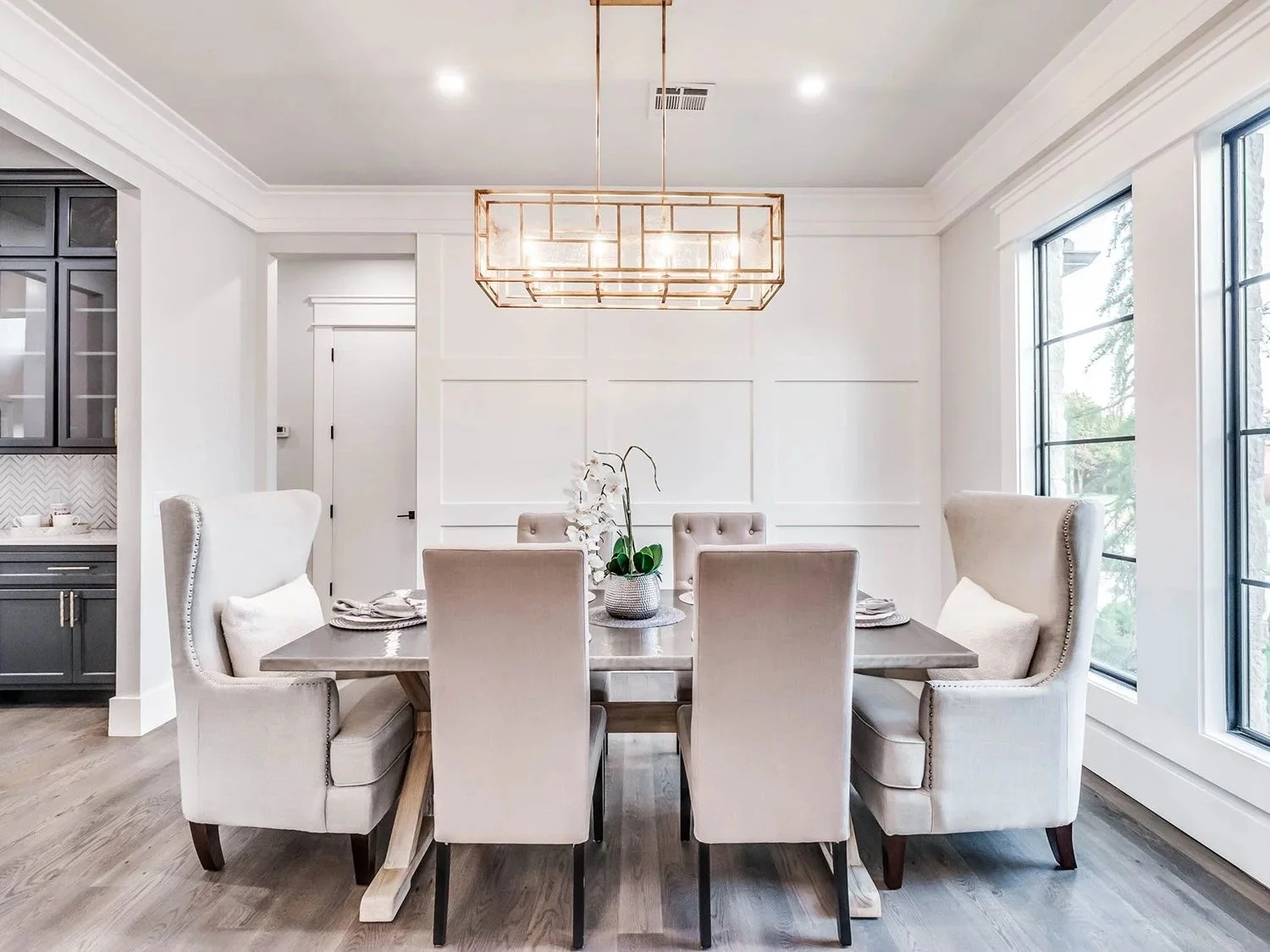 Dining room with a dark wood table, six beige upholstered chairs, a modern rectangular gold chandelier, white paneled wall, large windows, hardwood flooring, and a modern kitchen visible on the left.