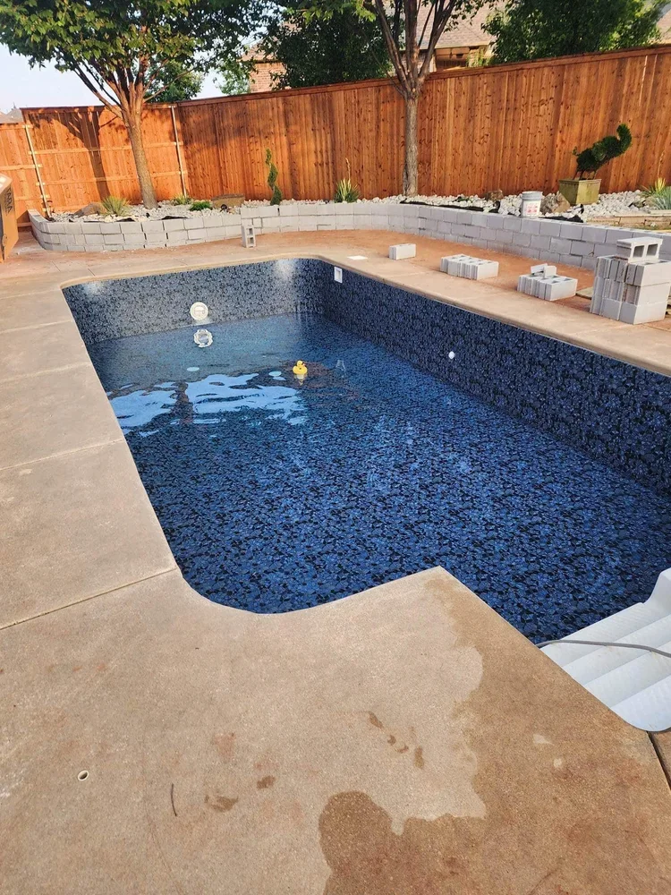 An empty swimming pool with some floating toys, surrounded by a concrete deck, with a fence and trees in the background, and construction materials on the patio.