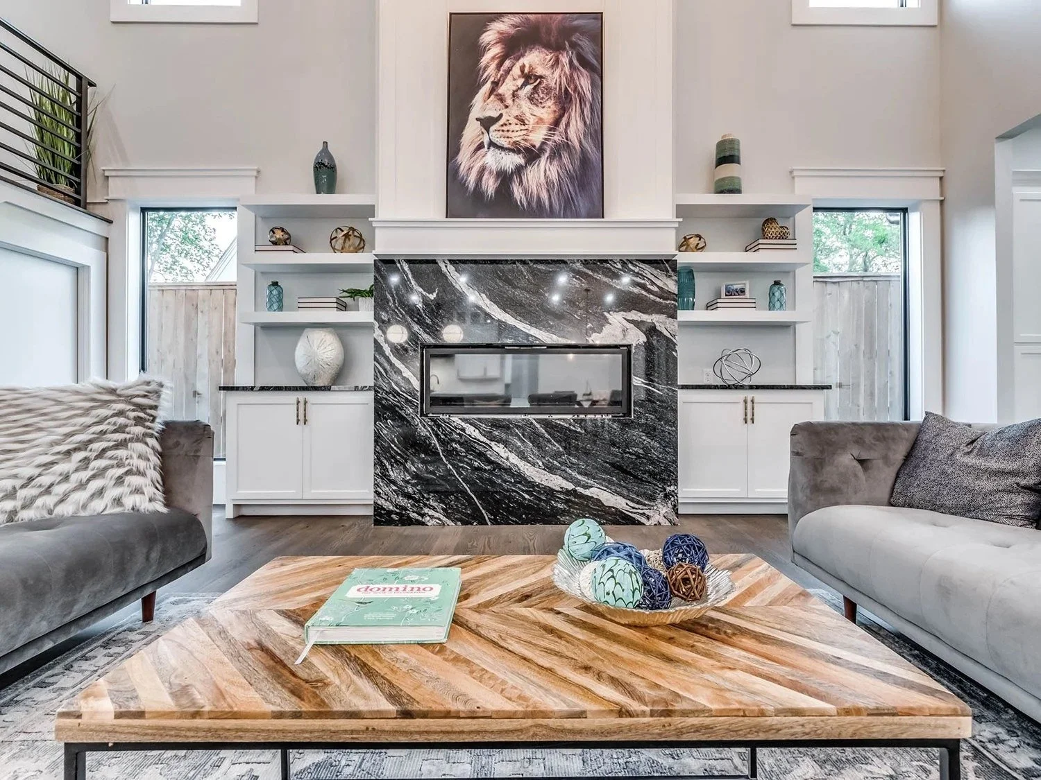 Living room with a centered black marble fireplace, white built-in shelves, gray sofas, a wooden coffee table with a decorative bowl of spherical objects, and a large lion portrait hanging above the fireplace.