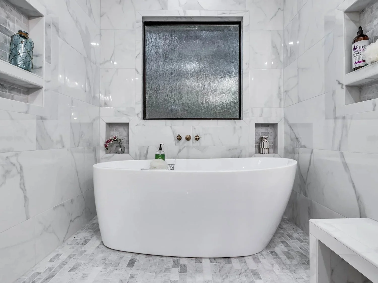 Modern bathroom with a white freestanding bathtub, marble tiled walls, built-in shelves with decorative items, and a small frosted window.