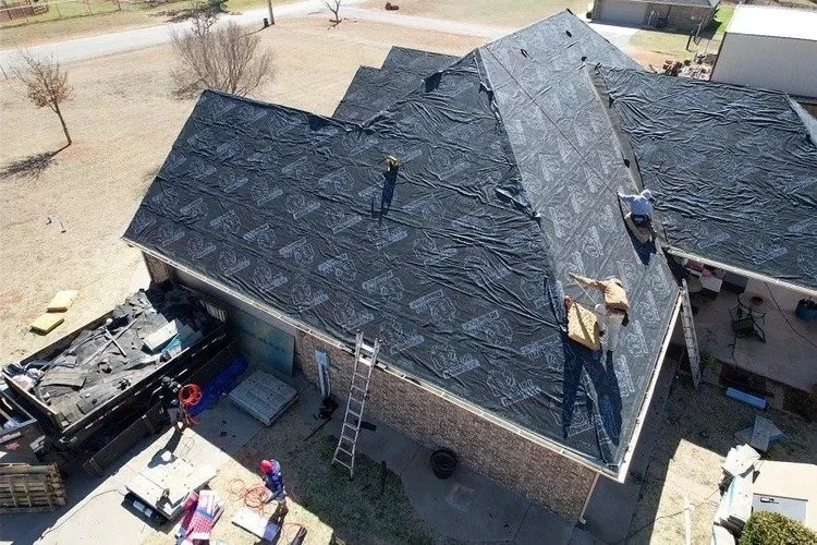 A house under construction with workers installing a new black shingle roof, ladders, and construction materials surrounding the area.
