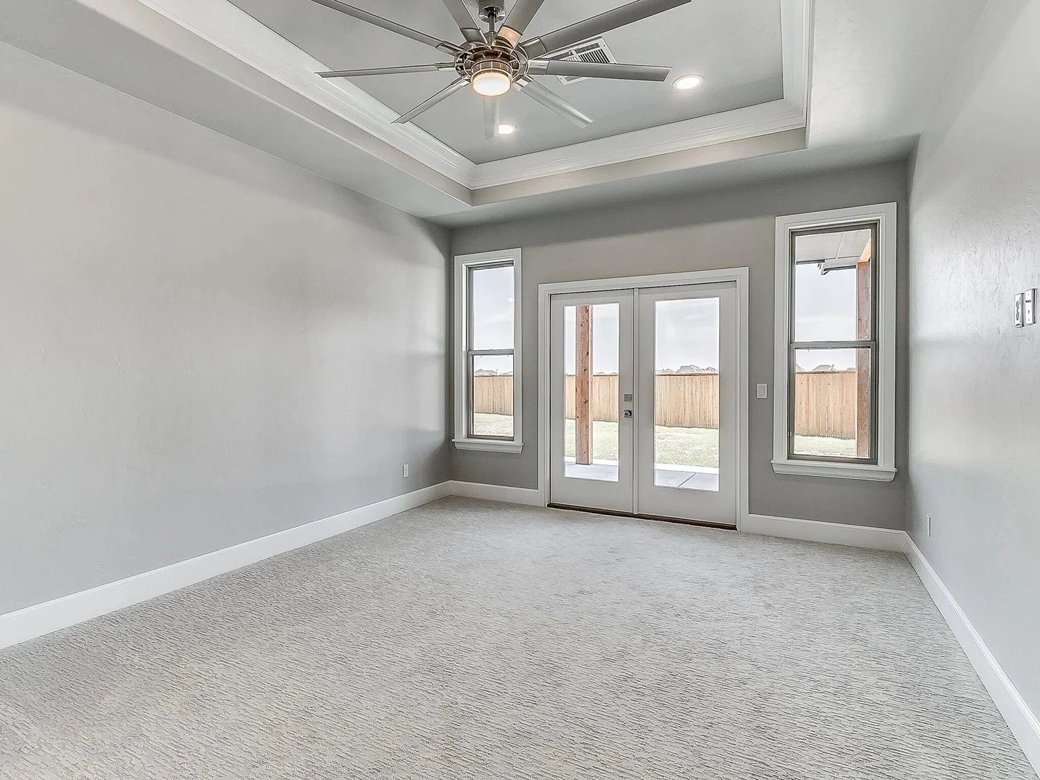 Empty room with light-colored walls, beige carpet, large windows, and a glass door leading outside. Ceiling has a tray design with a modern ceiling fan and recessed lighting.