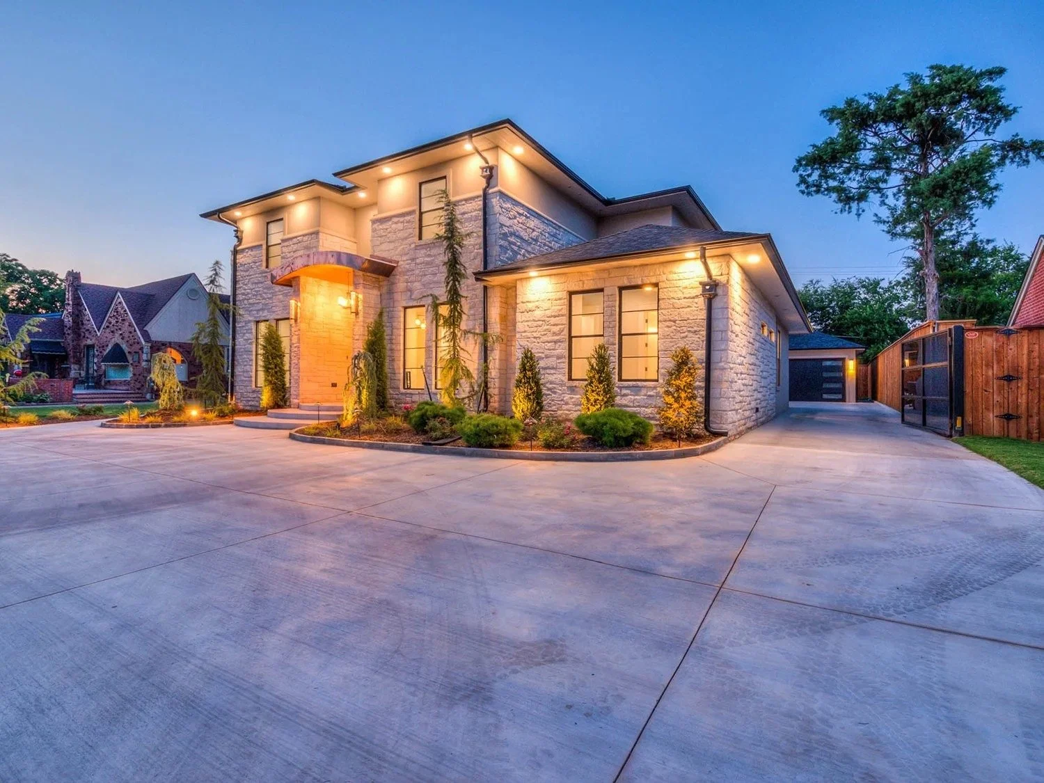 Modern two-story house with stone exterior and large windows, illuminated at dusk. Front yard landscaped with small bushes, trees, and pathway, with a concrete driveway and wooden fence.