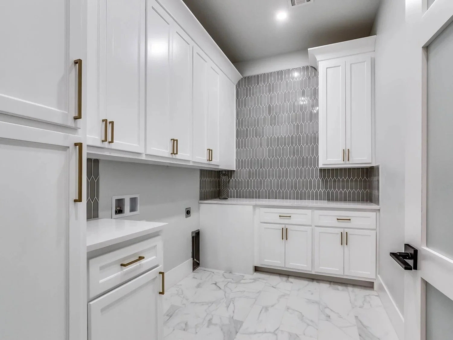 Modern laundry room with white cabinets, gold handles, marble floor, and a gray tile accent wall.