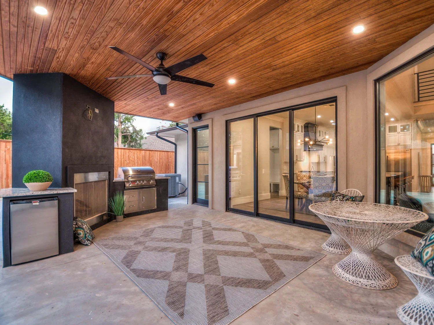 Outdoor covered patio with ceiling fan, recessed lighting, a rug, a small grill, a mini fridge, a potted plant, a cushioned chair with pillows, a glass-top table, and sliding glass doors leading inside a house.