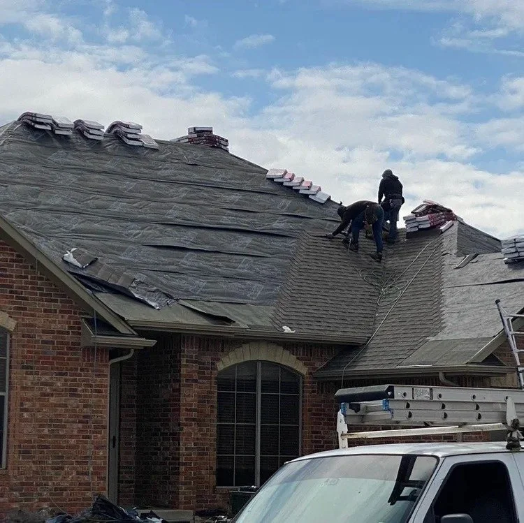Two workers on a sloped roof replacing or repairing shingles, with a truck parked nearby and a ladder leaning against the house.