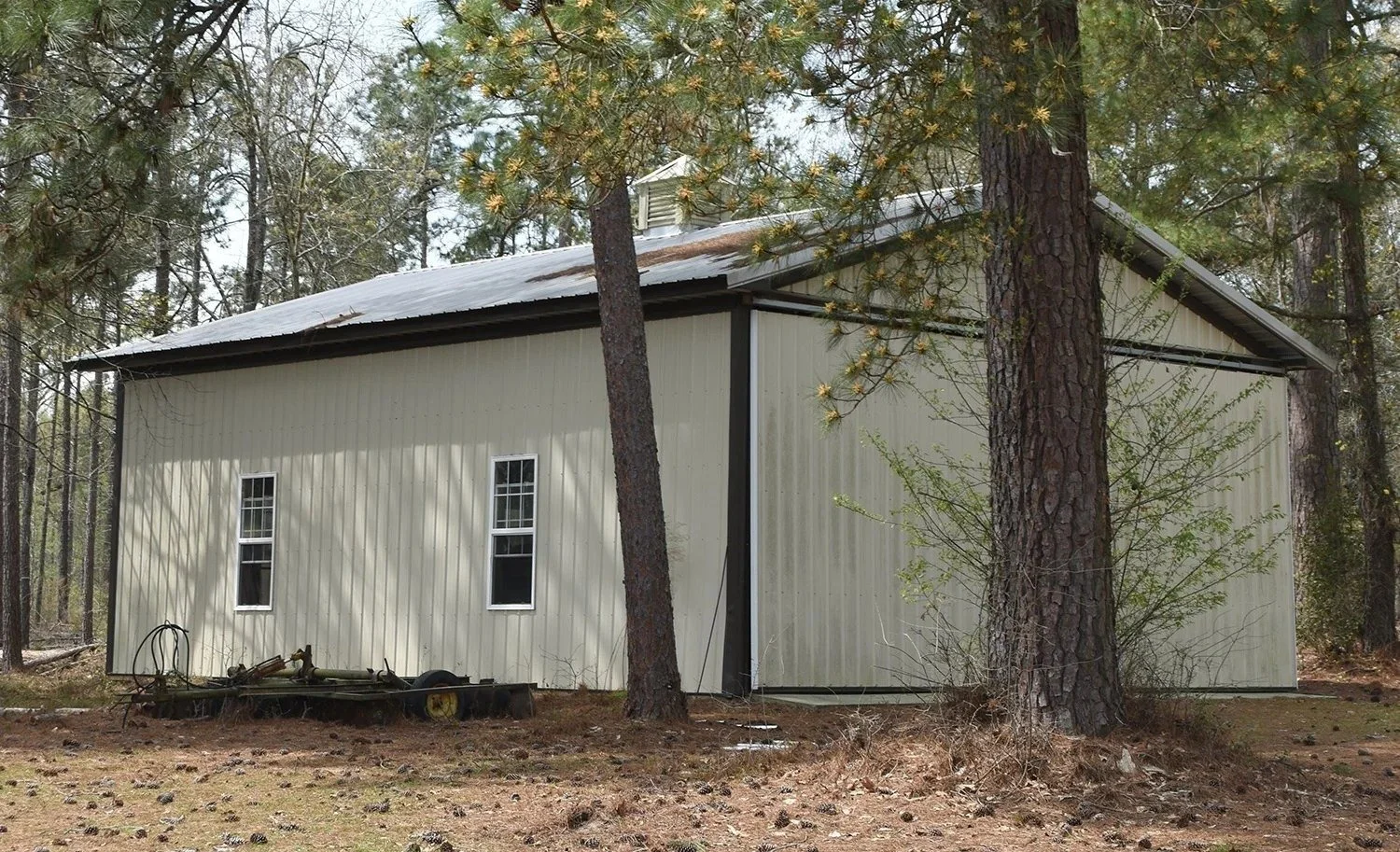 A beige metal building in a wooded area with a metal roof and two small windows, surrounded by pine trees.