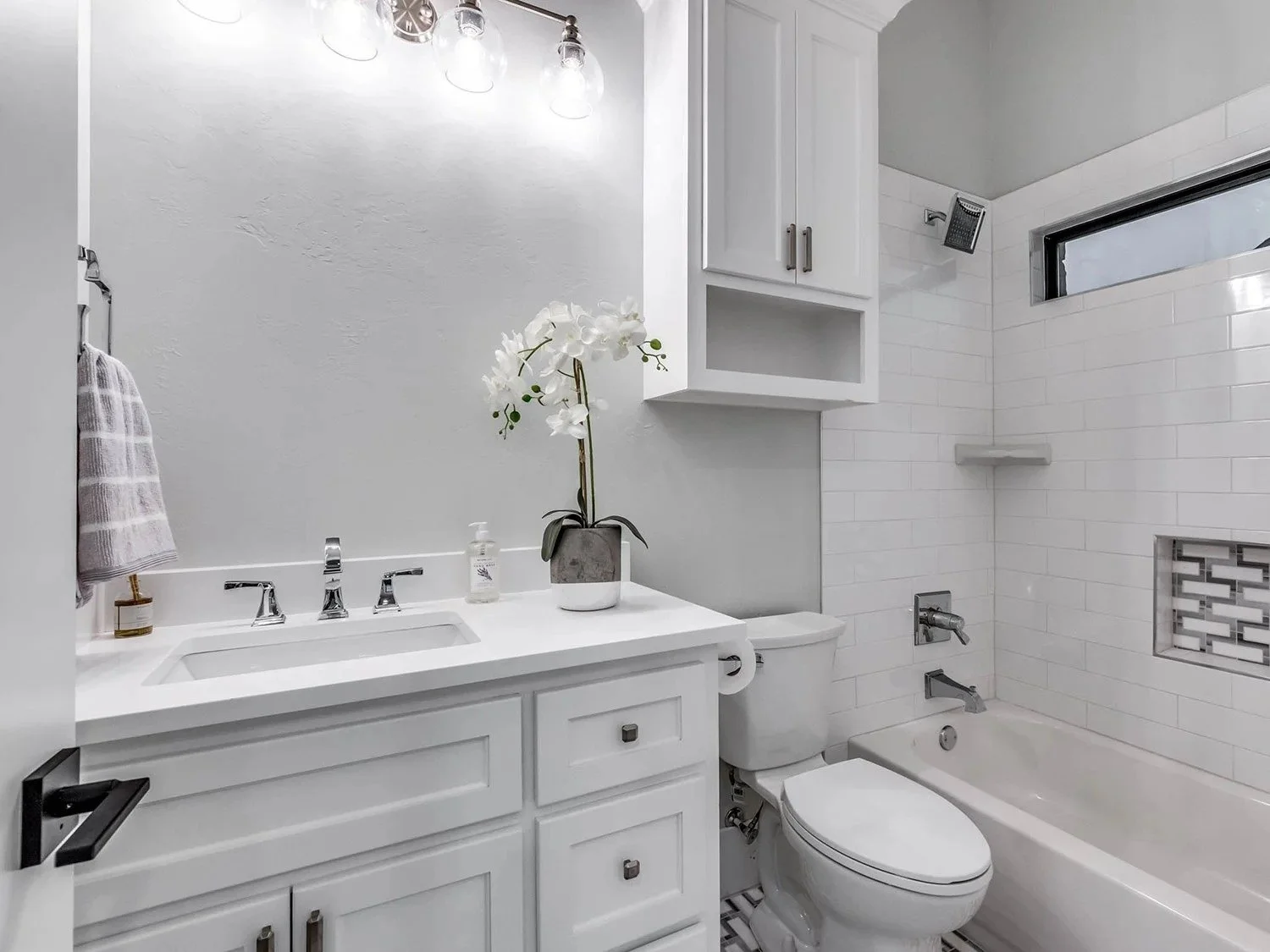 Modern white bathroom with a vanity, sink, and faucet, decorated with a potted white orchid, towel, soap dispenser, toilet, bathtub with showerhead, small window, and built-in shower shelf with a tile design.