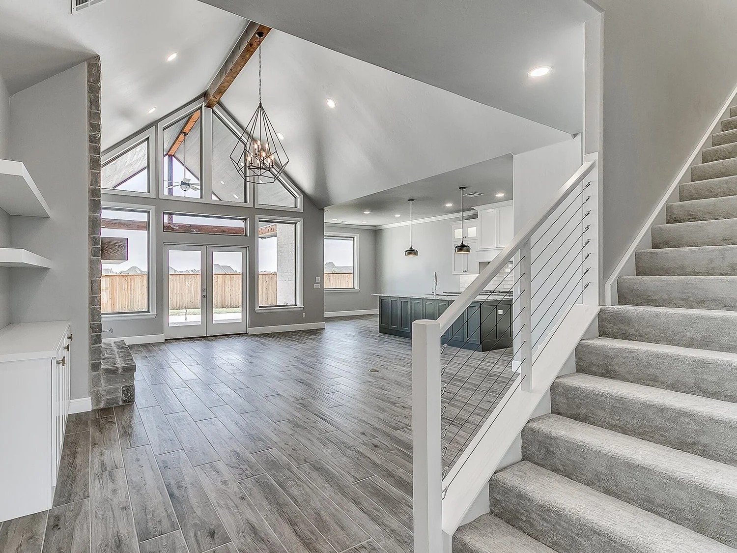 Open-concept living space with large windows, wood flooring, a staircase with a gray carpet, a kitchen with a black island, and modern light fixtures.