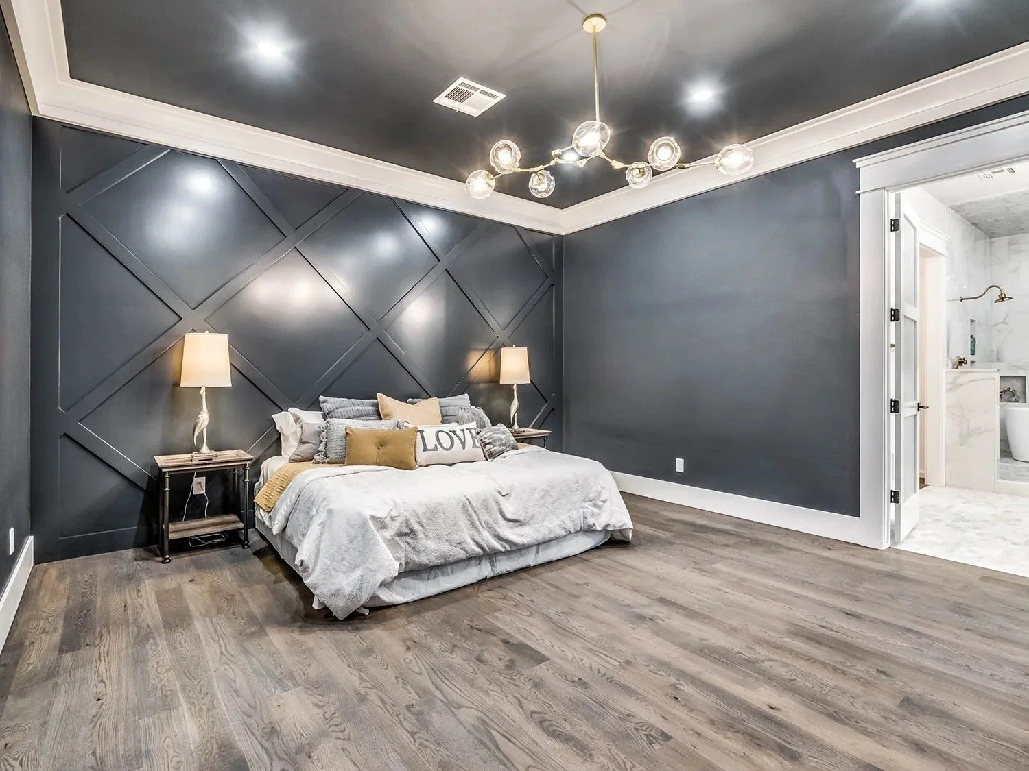 A modern bedroom with dark gray paneled accent wall, a bed with various gray and beige pillows, two bedside tables with lamps, and a chandelier with multiple glass globes. An open door leads to a bathroom with marble walls.