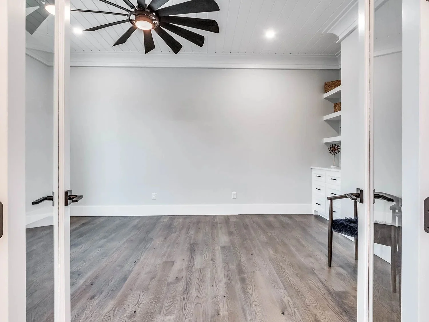 Empty room with white walls, gray hardwood floor, ceiling fan with black blades, built-in white shelves with baskets and decorative item, and a black furry chair with wooden legs near the right opening.