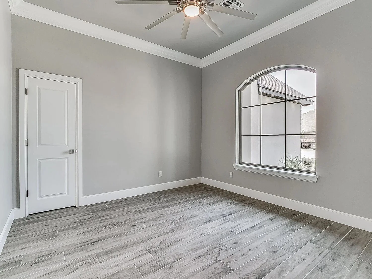 Empty room with gray walls, wood-style tile flooring, white baseboards, a large window, a door, and a ceiling fan.
