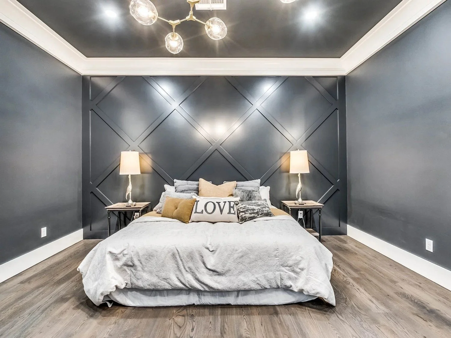 A modern bedroom with a dark gray wall featuring a geometric pattern, a white bed with gray, white, and beige pillows, and two nightstands with beige lamps. There is a contemporary chandelier with globe bulbs hanging from the ceiling.